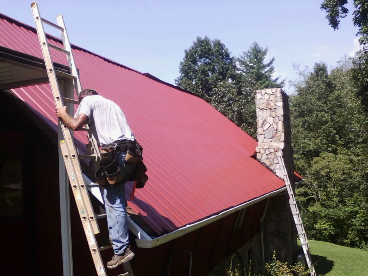 Roofer on a ladder beside a red metal roof and stone chimney — Soffit & Fascia Repair by Roofing Brothers in Phillipsburg